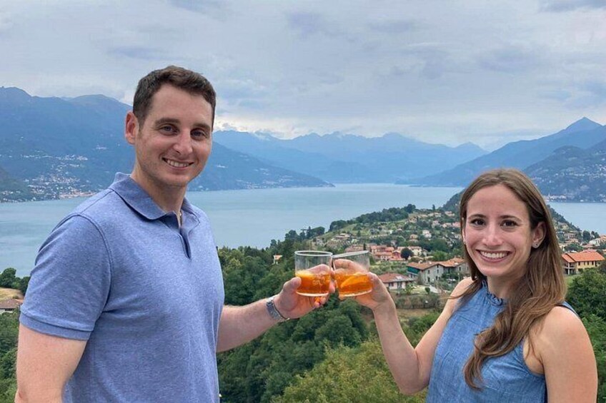 Twee personen proosten met drankjes, een meer, bergen en een stad op de achtergrond. 3