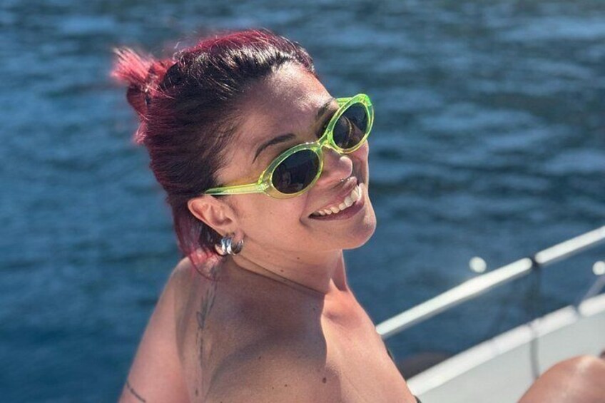 A person with red hair and green sunglasses, wearing a tattoo on their shoulder, standing by a body of water. 2