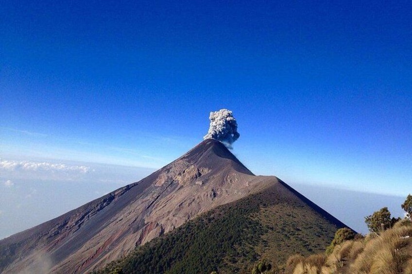 A volcano with a plume of smoke at its peak, surrounded by a forested landscape. 3