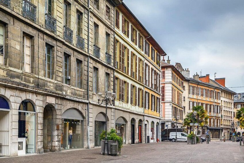 A historic European street with multi-story buildings, arched storefronts, and a paved plaza. 3
