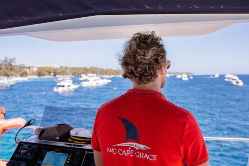 A person in a red shirt with 'ANC CAPE GRACE' on the back, steering a boat with a view of the sea and boats in the distance. 3