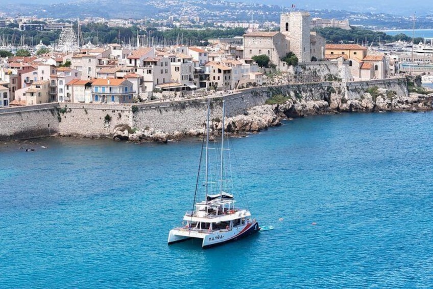 A sailboat named 'MAV' anchored near a coastal town with historic buildings and a stone wall. 3