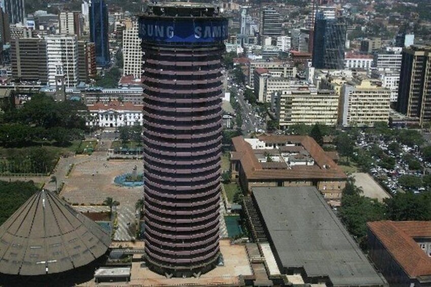 A tall building with a blue sign, surrounded by other buildings and a park. 4