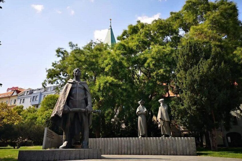 Una estatua de un hombre con capa, la aguja de una iglesia y árboles al fondo. 3