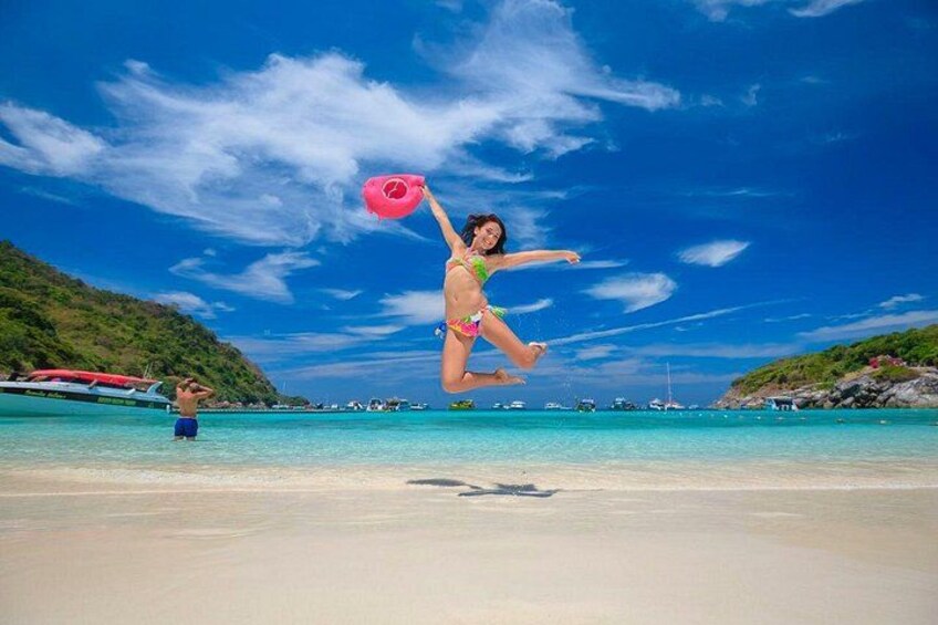 Una persona salta en la playa con un aro inflable rosa, cielo azul despejado y barcos en el agua. 1