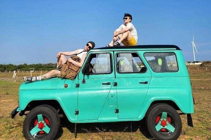 Un'auto d'epoca turchese con cerchi rossi parcheggiata in un campo, con una turbina eolica sullo sfondo. 2