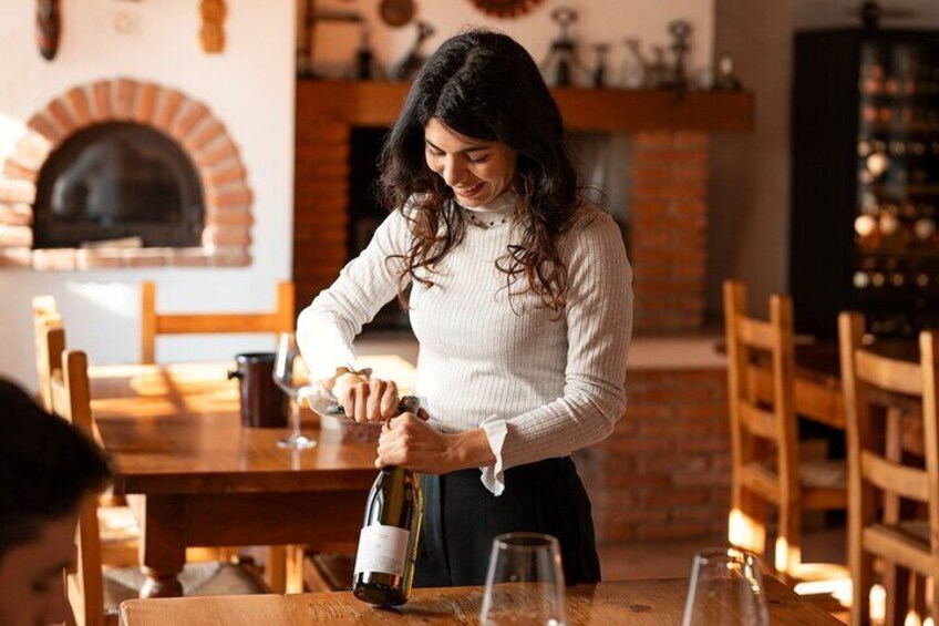 Une femme verse du vin dans un verre, assise à une table en bois dans un intérieur chaleureux, avec une cheminée en briques. 1