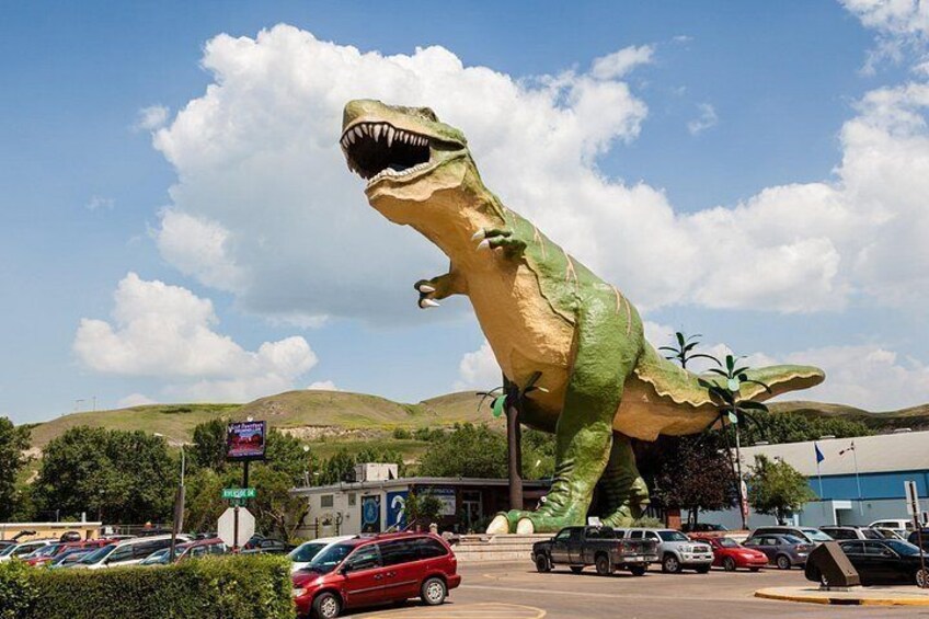 A large dinosaur statue in a parking lot with cars and a building in the background. 5