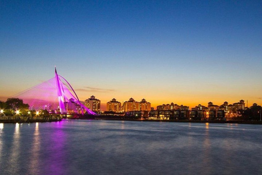 A cityscape at dusk with a prominent bridge illuminated in purple, reflecting on the water. 1