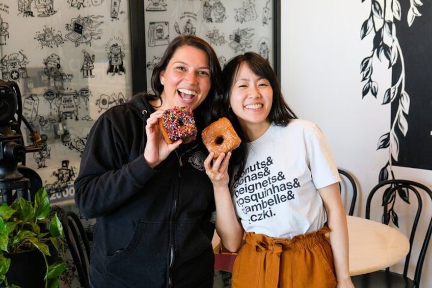 Two people holding donuts in a room with a wall decorated with drawings. 1