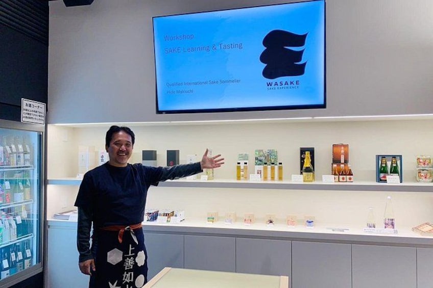 En person står framför en utställning med olika produkter, och ovanför finns en skärm som visar texten ”Workshop: Lär dig om och provsmaka sake”. 4