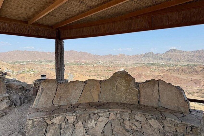 A stone bench under a wooden shelter with a view of a desert landscape and mountains. 2