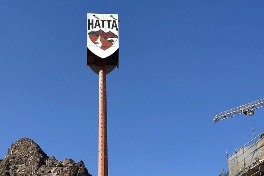A sign with the word 'HATTA' and a mountain graphic, a tall pole, and a construction crane against a clear blue sky. 5
