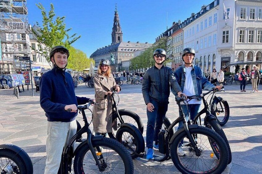 Three people standing with bicycles in a public square with buildings and a tower in the background. 1