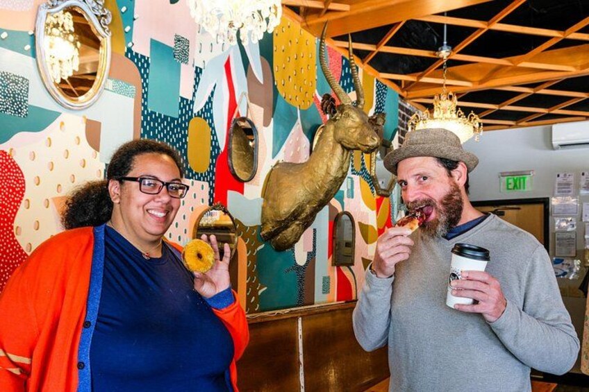 Two people holding donuts and coffee cups in a room with colorful abstract art and a large deer sculpture. 2