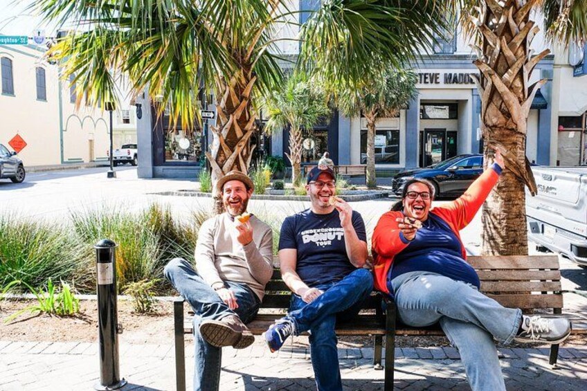 Three people sitting on a bench in a sunny outdoor setting with palm trees and a building in the background. 4