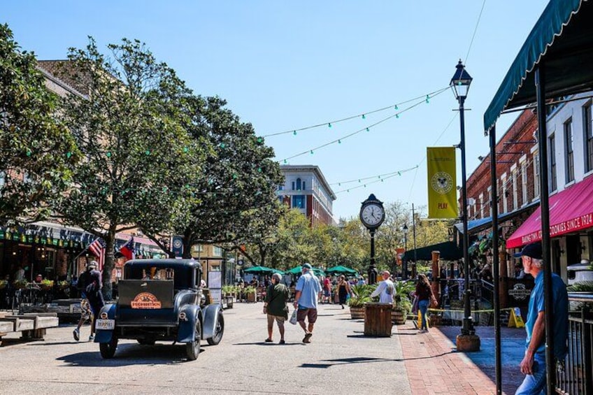 A bustling street with pedestrians, vintage cars, and outdoor seating at cafes. 3