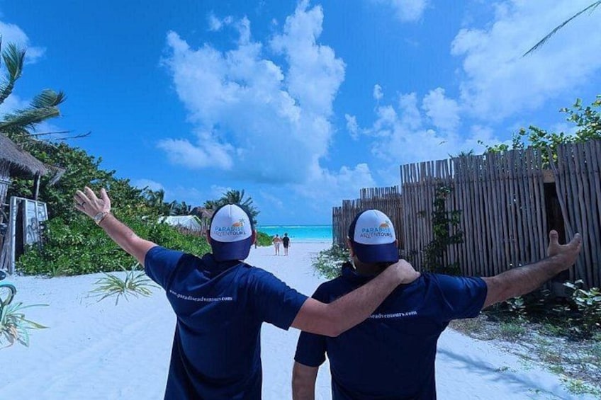 Two people wearing matching blue shirts and hats, standing on a sandy beach with a wooden fence and palm trees in the background. 3