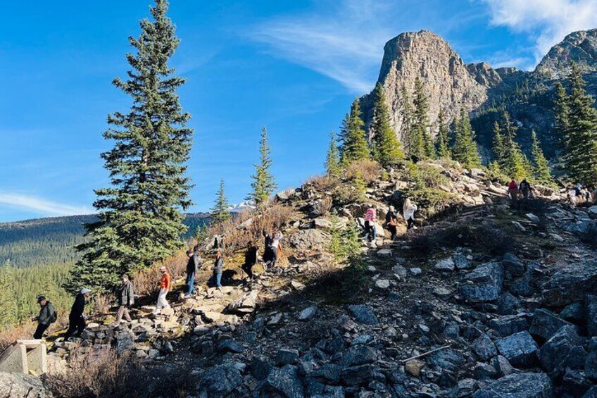 Un sentier de montagne rocailleux, avec des randonneurs, de grands arbres à feuilles persistantes et un pic rocheux imposant en arrière-plan. 2