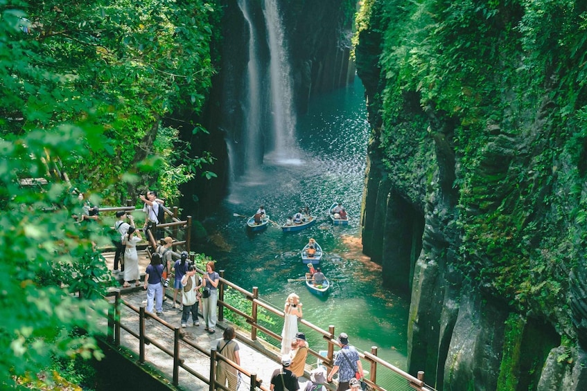 A waterfall in a lush green canyon with people on a bridge and canoes on the water. 1