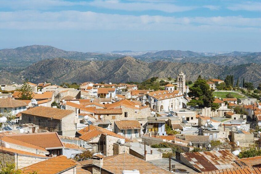 A town with terracotta-roofed buildings, a prominent church with a bell tower, and a mountainous backdrop. 3