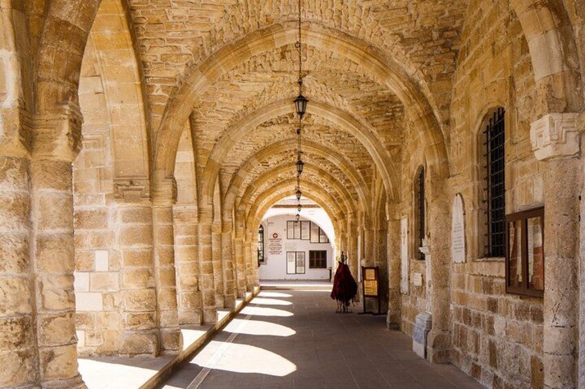A stone corridor with arches, a hanging light fixture, and a distant building. 4