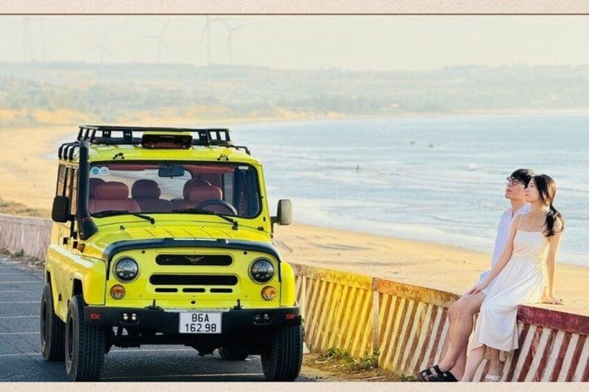 Ein gelbes Geländefahrzeug steht auf einem Strand, neben der Uferbefestigung stehen ein Paar. 3