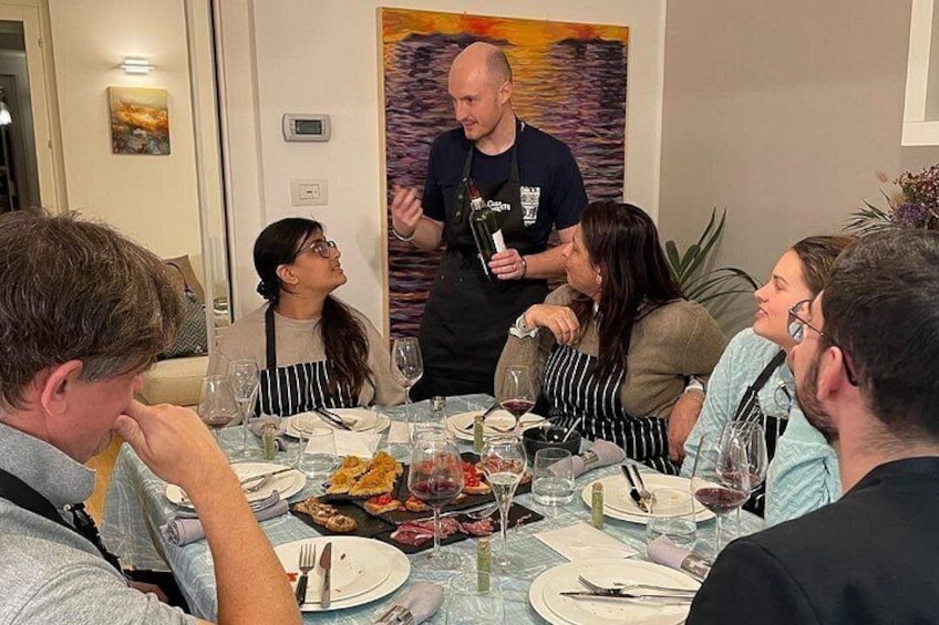 Un grupo de personas cenando en una mesa, con copas de vino, platos y un camarero. 2