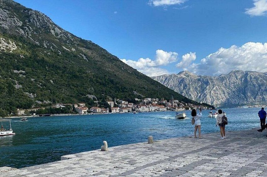 Un pueblo costero con un muelle de piedra, gente de pie junto al agua, barcos y montañas al fondo. 4