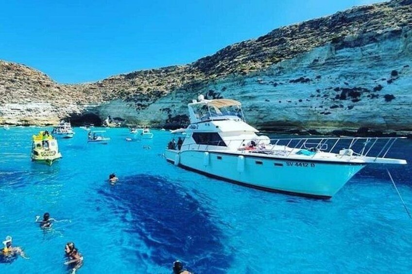 Una barca bianca con la targa SV 4412 D su un mare blu intenso, circondata da imbarcazioni più piccole e persone che nuotano. 1