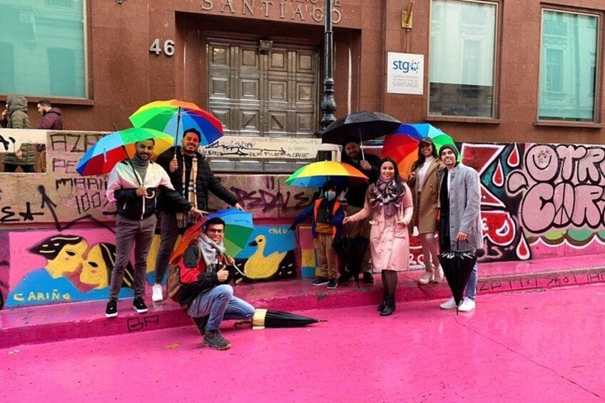 Un groupe de personnes posant avec des parapluies colorés devant un mur couvert de graffitis, avec un bâtiment en arrière-plan. 3