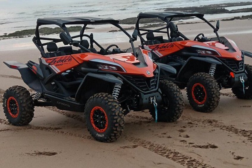 Two orange and black off-road vehicles parked on a sandy beach with the ocean in the background. 4