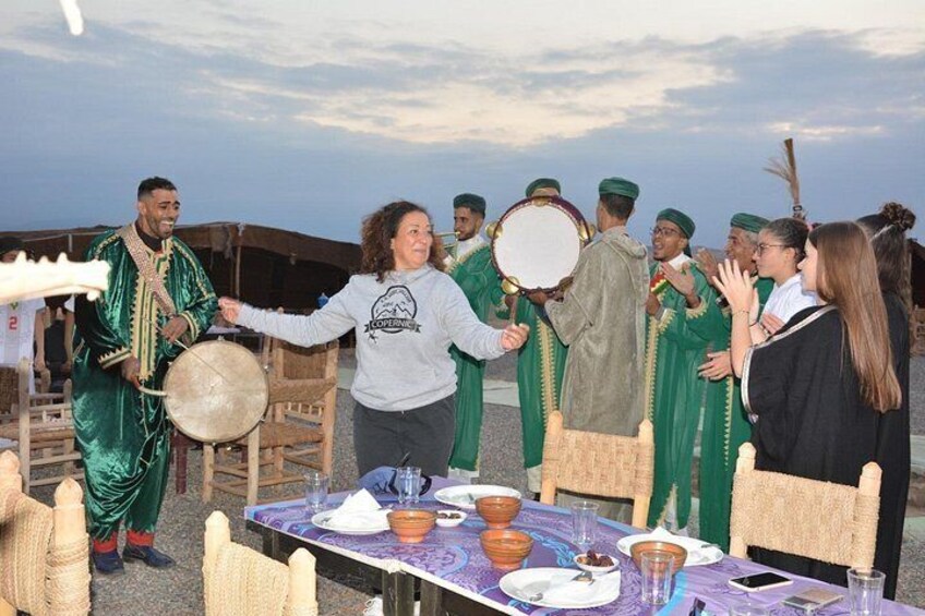 Un groupe de personnes est assis autour d’une table, devant des plats traditionnels. On y voit également un joueur de tambour et des personnes vêtues de vert. 4