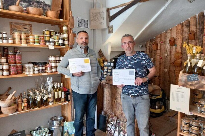 Two men holding certificates in a rustic shop with shelves of jars and bottles. 4