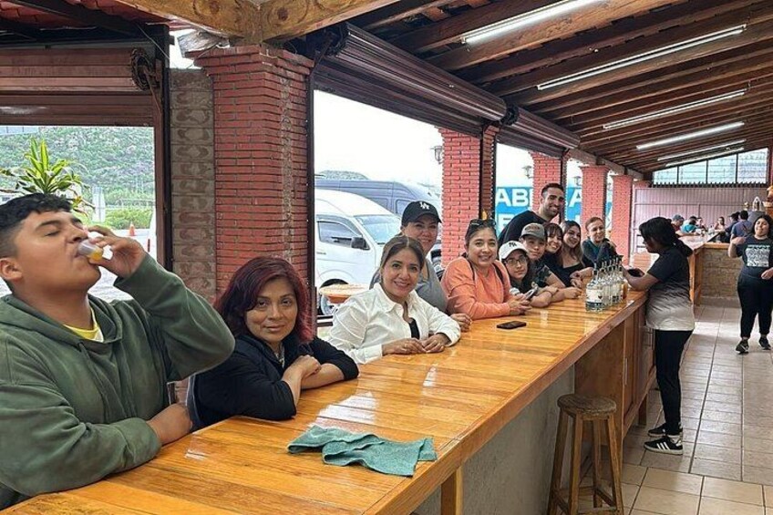 A group of people sitting at a wooden counter in a restaurant with brick pillars and a tiled floor. 2