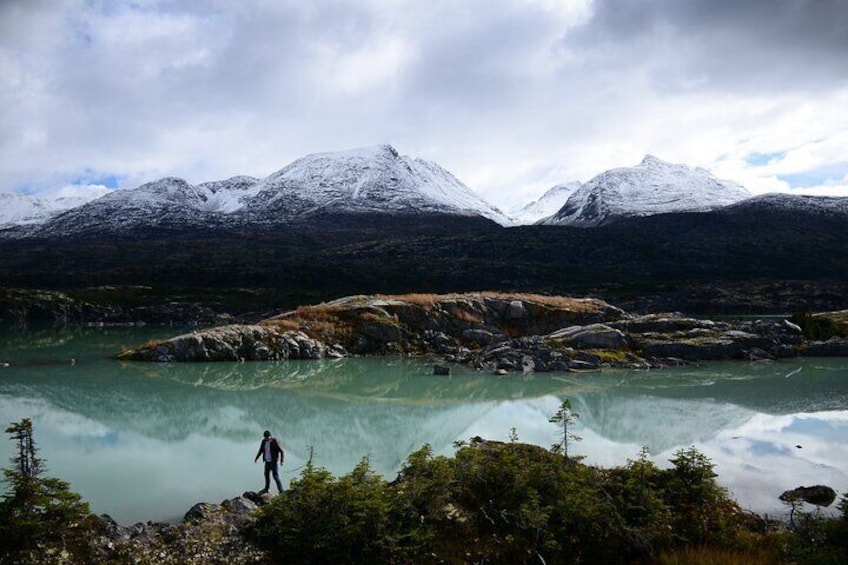A person standing on a rocky outcrop near a turquoise lake with snow-capped mountains in the background. 4