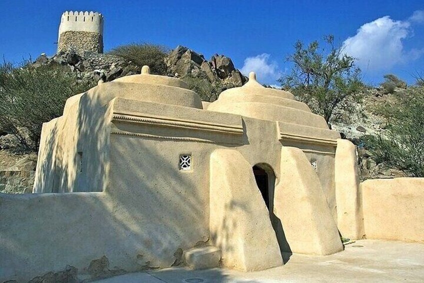 A large, beige, fortress-like structure with a central archway, set against a rocky hillside under a clear blue sky. 2