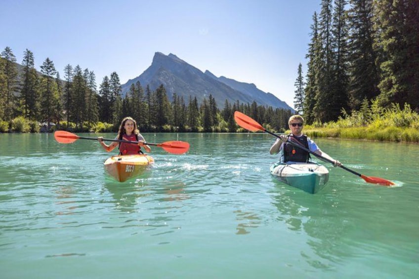 Two people kayaking on a calm river surrounded by forested mountains. 1