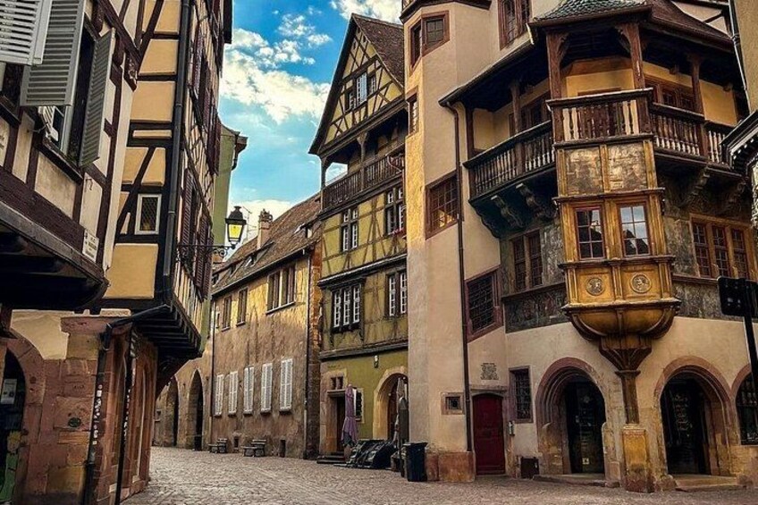 Una calle empedrada bordeada por edificios tradicionales con entramado de madera, una puerta roja y una torre del reloj. 2