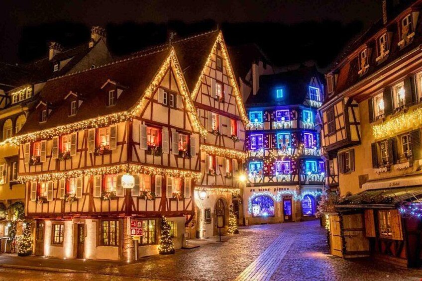 Una plaza histórica en una ciudad europea, iluminada con luces de colores, con edificios tradicionales de entramado de madera y calles adoquinadas. 4