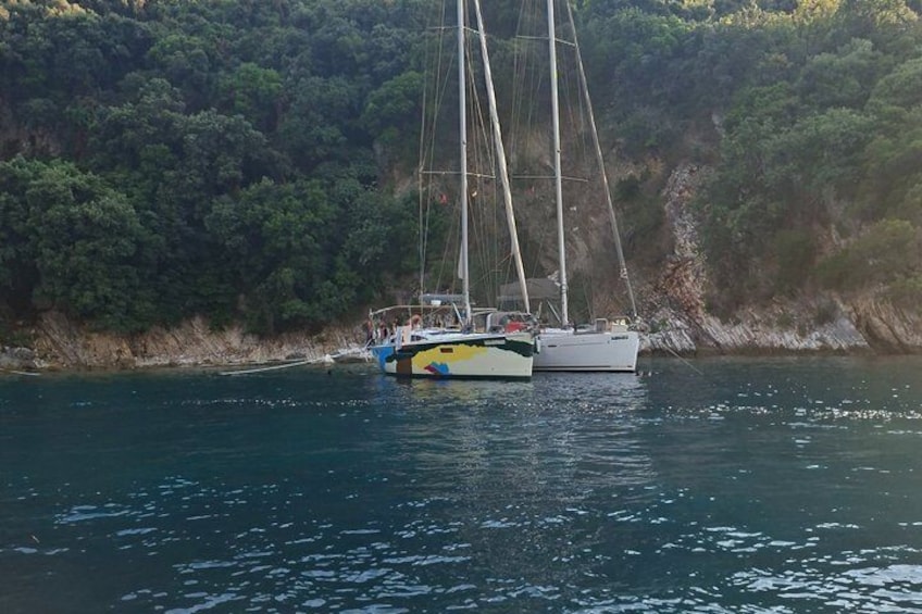 A sailboat with a cabin and a smaller boat docked near a rocky shoreline with dense greenery. 1