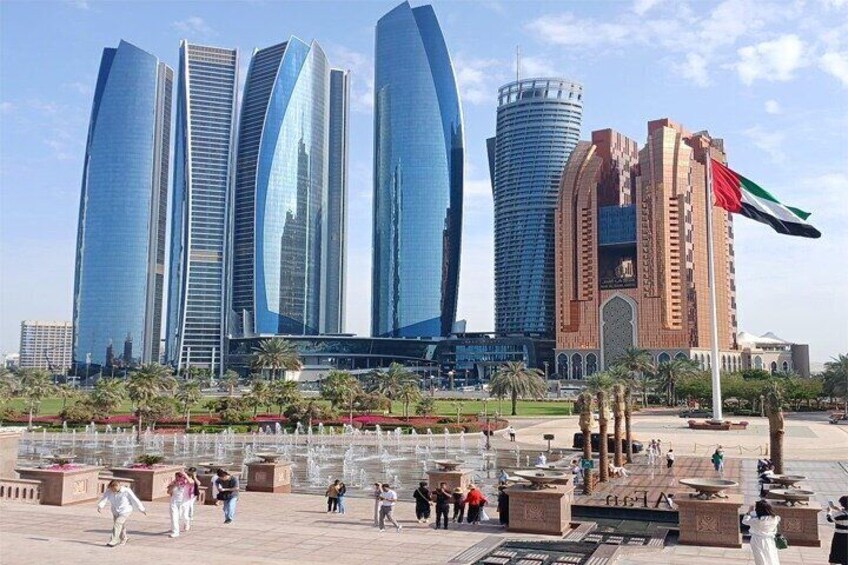 A modern cityscape with tall buildings, a central fountain, and people walking. 1