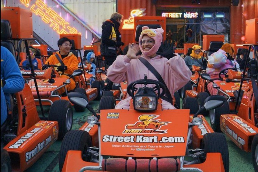 Une personne vêtue d’un costume rose conduit un petit véhicule orange équipé d’un volant et d’une ceinture de sécurité, et est entourée de véhicules similaires ainsi que de personnes portant des costumes orange. 4