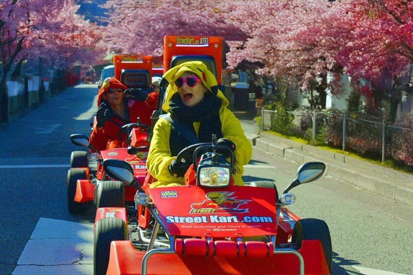 A street kart race with participants in yellow and red costumes, a red kart, and cherry blossom trees lining the street. 2