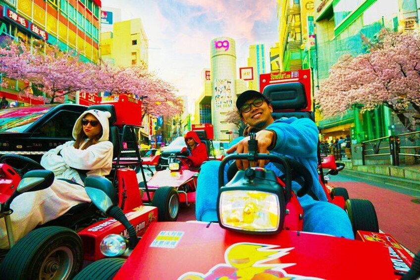A red vehicle with a person driving, another person sitting, and cherry blossom trees lining the street. 1
