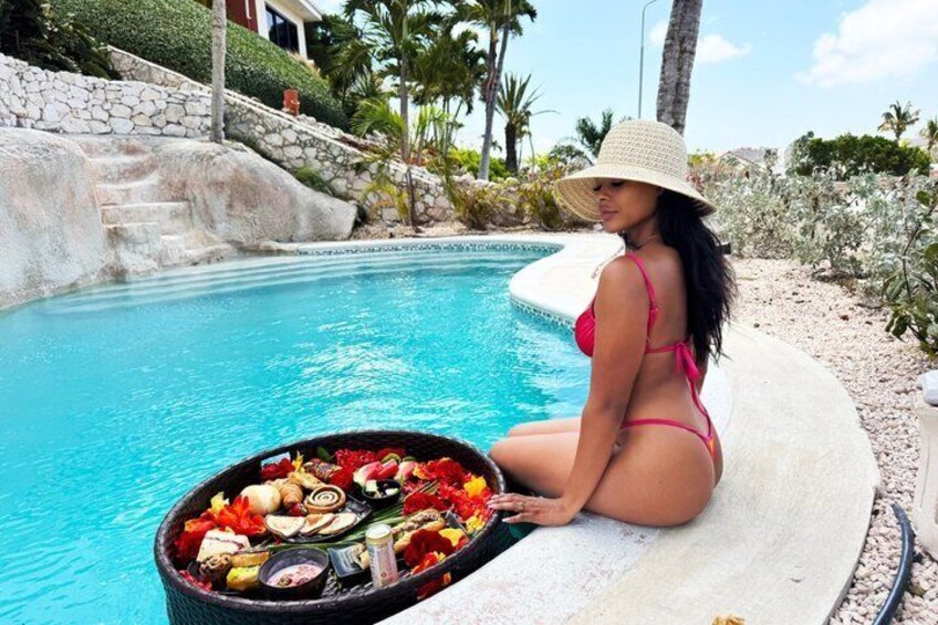 A woman in a bikini sitting by a pool with a basket of food and drinks. 2