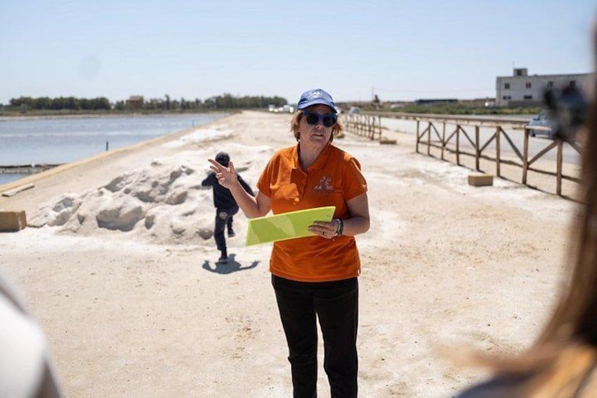Une personne portant un t-shirt orange et une casquette bleue, tenant un dossier jaune sur une plage de sable, avec une clôture en bois et une étendue d'eau en arrière-plan. 3