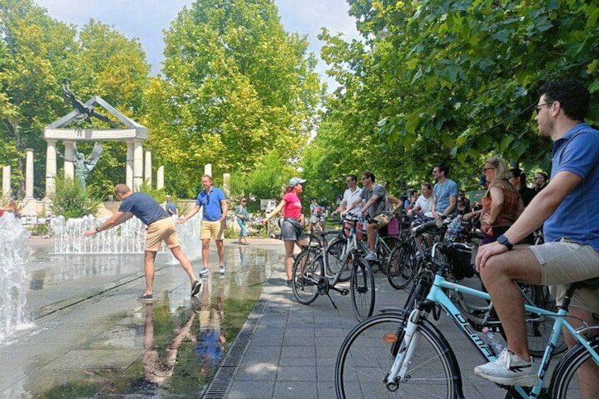 Personas montando en bicicleta e interactuando con una fuente en un parque. 2
