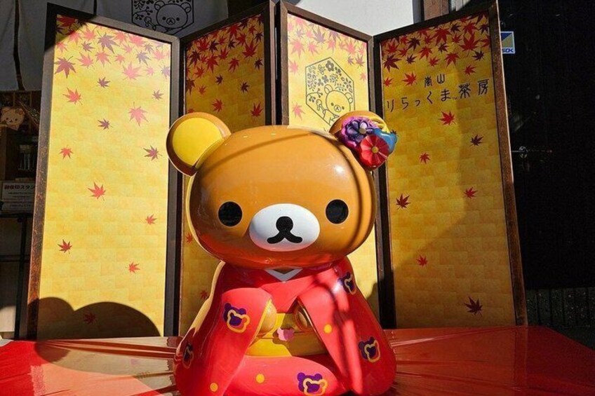 A large teddy bear in a red outfit with a bow, standing in front of a traditional Japanese sliding door with autumn leaves and a decorative emblem. 5
