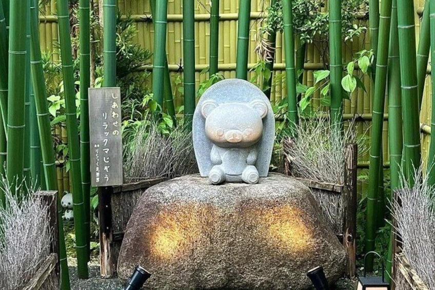 A stone sculpture of a bear sitting on a rock, surrounded by bamboo plants and a wooden sign with Japanese characters. 3